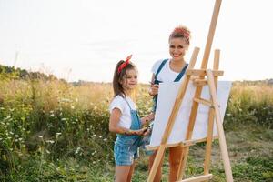 beleza mãe pintura com dela pequeno filha. à moda mulher desenhando a cenário com pequeno garota. fofa criança dentro uma branco camiseta e azul jeans. mãe dia. foto