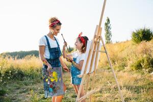 beleza mãe pintura com dela pequeno filha. à moda mulher desenhando a cenário com pequeno garota. fofa criança dentro uma branco camiseta e azul jeans. mãe dia. foto