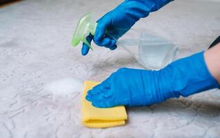 pessoas, tarefas domésticas e serviço de limpeza conceito - fechar acima do mulher dentro borracha luvas com pano e derergente spray limpeza tapete às lar. fechar acima do mulher com pano limpeza tapete foto