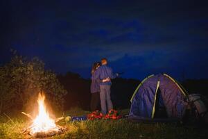 amoroso casal caminhantes desfrutando cada outro, em pé de fogueira às noite debaixo tarde céu perto árvores e barraca. romântico acampamento perto floresta dentro a montanhas foto