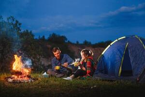 acampamento noite dentro montanhas. turista casal sentado dentro frente do iluminado barraca aceso de queimando fogueira. turismo e ao ar livre atividade conceito. foto