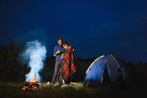 amoroso casal caminhantes desfrutando cada outro, em pé de fogueira às noite debaixo tarde céu perto árvores e barraca. romântico acampamento perto floresta dentro a montanhas foto