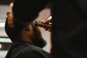 retrato do jovem Preto homem ser aparado com profissional elétrico tosquiadeira máquina dentro barbearia.masculino beleza tratamento conceito. jovem africano cara obtendo Novo corte de cabelo dentro barbeiro salão foto