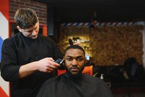 retrato do jovem Preto homem ser aparado com profissional elétrico tosquiadeira máquina dentro barbearia.masculino beleza tratamento conceito. jovem africano cara obtendo Novo corte de cabelo dentro barbeiro salão foto