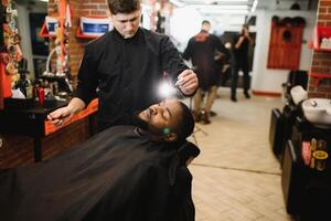 lado Visão do sério homem com à moda moderno corte de cabelo olhando frente dentro barbeiro fazer compras. mão do barbeiro guardando em linha reta navalha e corte na moda listras em cabeça do cliente. foto