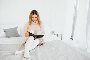 uma mulher lendo uma livro e sorridente Como ela senta dentro cama. a alarme relógio em a escrivaninha ao lado dela. foto