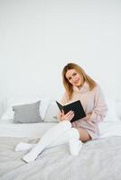 uma mulher lendo uma livro e sorridente Como ela senta dentro cama. a alarme relógio em a escrivaninha ao lado dela. foto