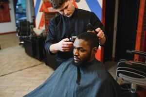 retrato do jovem Preto homem ser aparado com profissional elétrico tosquiadeira máquina dentro barbearia.masculino beleza tratamento conceito. jovem africano cara obtendo Novo corte de cabelo dentro barbeiro salão foto