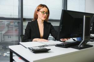 bonito, legal, bonitinho, perfeito mulher sentado às dela escrivaninha em couro cadeira dentro trabalhos estação, vestindo copos, roupa formal, tendo computador portátil e caderno em a mesa foto