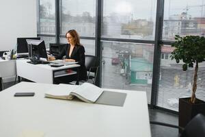 bonito, legal, bonitinho, perfeito mulher sentado às dela escrivaninha em couro cadeira dentro trabalhos estação, vestindo copos, roupa formal, tendo computador portátil e caderno em a mesa foto