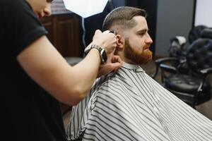 jovem bonito barbeiro fazer corte de cabelo do atraente barbudo homem dentro barbearia. foto