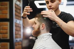 jovem bonito barbeiro fazer corte de cabelo do atraente barbudo homem dentro barbearia. foto