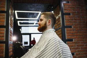 fazer corte de cabelo Veja perfeito. jovem barbudo homem obtendo corte de cabelo de cabeleireiro enquanto sentado dentro cadeira às barbearia. foto