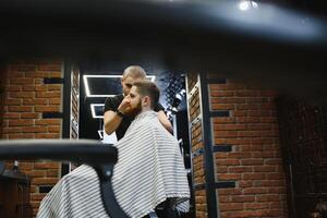 jovem bonito barbeiro fazer corte de cabelo do atraente barbudo homem dentro barbearia. foto