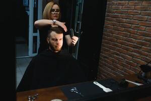 servindo cliente dentro barbearia. profissional barbeiro garota, fêmea cabeleireiro fazer moderno corte de cabelo para uma homem sentado dentro barbeiro fazer compras cadeira. foco em uma garota. cabeleireiro, barbear, corte, asseio. foto