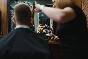 à moda homem sentado barbeiro fazer compras estilista de cabelo cabeleireiro mulher corte dele cabelo retrato bonito feliz jovem barbudo caucasiano cara obtendo na moda corte de cabelo atraente barbeiro menina trabalhando servindo cliente. foto