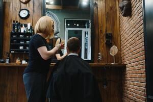 servindo cliente dentro barbearia. profissional barbeiro garota, fêmea cabeleireiro fazer moderno corte de cabelo para uma homem sentado dentro barbeiro fazer compras cadeira. foco em uma garota. cabeleireiro, barbear, corte, asseio. foto