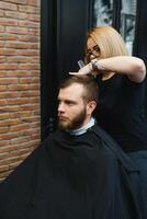 servindo cliente dentro barbearia. profissional barbeiro garota, fêmea cabeleireiro fazer moderno corte de cabelo para uma homem sentado dentro barbeiro fazer compras cadeira. foco em uma garota. cabeleireiro, barbear, corte, asseio. foto