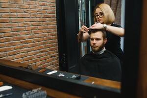 servindo cliente dentro barbearia. profissional barbeiro garota, fêmea cabeleireiro fazer moderno corte de cabelo para uma homem sentado dentro barbeiro fazer compras cadeira. foco em uma garota. cabeleireiro, barbear, corte, asseio. foto