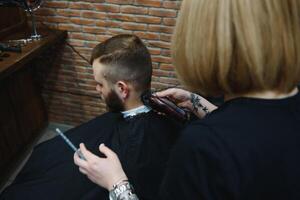 à moda homem sentado barbeiro fazer compras estilista de cabelo cabeleireiro mulher corte dele cabelo retrato bonito feliz jovem barbudo caucasiano cara obtendo na moda corte de cabelo atraente barbeiro menina trabalhando servindo cliente. foto