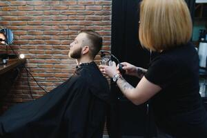 servindo cliente dentro barbearia. profissional barbeiro garota, fêmea cabeleireiro fazer moderno corte de cabelo para uma homem sentado dentro barbeiro fazer compras cadeira. foco em uma garota. cabeleireiro, barbear, corte, aliciamento foto