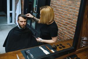 servindo cliente dentro barbearia. profissional barbeiro garota, fêmea cabeleireiro fazer moderno corte de cabelo para uma homem sentado dentro barbeiro fazer compras cadeira. foco em uma garota. cabeleireiro, barbear, corte, asseio. foto