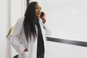 medicamento, tecnologia e cuidados de saúde conceito - feliz sorridente africano americano fêmea médico ou dentro branco casaco com estetoscópio chamando em Smartphone sobre hospital fundo. foto