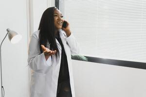 medicamento, tecnologia e cuidados de saúde conceito - feliz sorridente africano americano fêmea médico ou dentro branco casaco com estetoscópio chamando em Smartphone sobre hospital fundo. foto