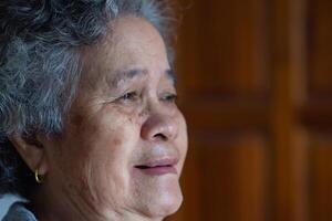 fechar-se do voltado para uma Senior mulher sorridente e olhando longe enquanto sentado em uma cadeira dentro uma vivo sala. conceito do envelhecido pessoas e cuidados de saúde foto