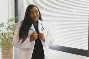 retrato do sorridente fêmea médico vestindo branco casaco com estetoscópio dentro hospital escritório. foto
