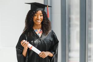 alegre afro americano fêmea graduado em pé dentro frente do universidade construção foto