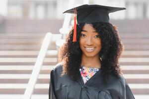 alegre afro americano fêmea graduado em pé dentro frente do universidade construção foto