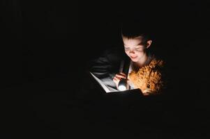 criança lendo livro dentro cama. crianças ler às noite. pequeno Garoto com fada conto livros dentro quarto . Educação para jovem crianças. hora de dormir história dentro a tarde. fofa criança debaixo cobertor dentro Sombrio quarto com luminária foto