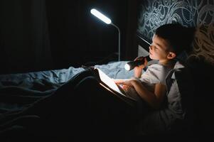 criança lendo livro dentro cama. crianças ler às noite. pequeno Garoto com fada conto livros dentro quarto . Educação para jovem crianças. hora de dormir história dentro a tarde. fofa criança debaixo cobertor dentro Sombrio quarto com luminária foto