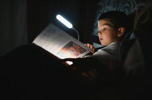 criança lendo livro dentro cama. crianças ler às noite. pequeno Garoto com fada conto livros dentro quarto . Educação para jovem crianças. hora de dormir história dentro a tarde. fofa criança debaixo cobertor dentro Sombrio quarto com luminária foto