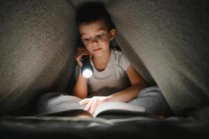 lendo livro e usando lanterna. menino em roupas casuais, deitado perto da barraca no entardecer foto