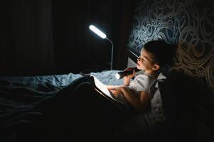 uma Garoto do 5-6 anos velho é lendo uma livro dentro a tarde dentro a Sombrio debaixo uma cobertor com uma brinquedo urso. foto