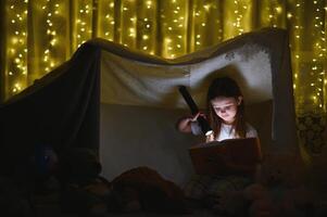 pequeno menina estão lendo uma livro com lanternas dentro barraca. feliz menina jogando às lar. engraçado adorável criança tendo Diversão dentro sala. foto