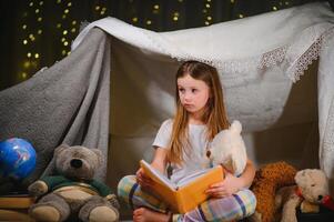 pequeno menina estão lendo uma livro com lanternas dentro barraca. feliz menina jogando às lar. engraçado adorável criança tendo Diversão dentro sala. foto