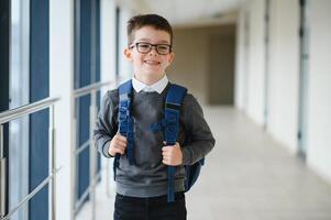 alegre sorridente pequeno Garoto com grande mochila tendo Diversão. escola conceito. costas para escola foto