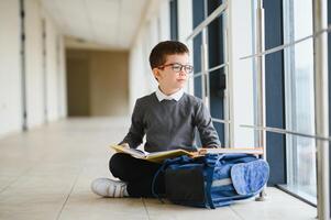 feliz fofa esperto Garoto dentro óculos com escola saco e livro dentro dele mão. primeiro Tempo para escola. costas para escola. foto