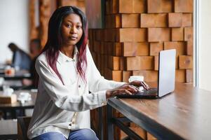 lindo africano o negócio mulher esperando para a encontro dentro uma co-trabalho espaço e sentado com a portátil computador. hipster menina é surfar a rede em uma computador portátil durante almoço. foto