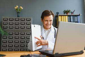 Tiros na Cabeça retrato inscrição tela Visão do meia idade fêmea médico ou gp conversa consultar cliente conectados em computador, mulher enfermeira falar com doente paciente em ligar usar Webcam conferência em computador portátil. foto