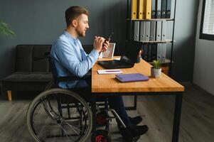 Desativado pessoa dentro a cadeira de rodas trabalho dentro a escritório às a computador. ele é sorridente e apaixonado sobre a fluxo de trabalho foto