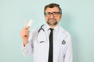 confiante masculino médico dentro branco médico Jaqueta segurando pílulas enquanto olhando às a Câmera isolado em azul fundo. remédio conceito. foto