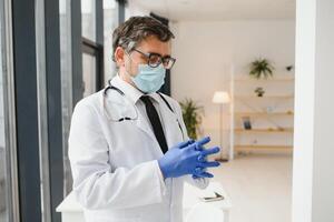 masculino médico dentro uma protetora mascarar coloca em luvas dentro a clínica foto
