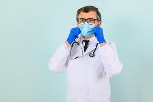 homem dentro médico luvas colocando em protetora face mascarar contra azul fundo. foto