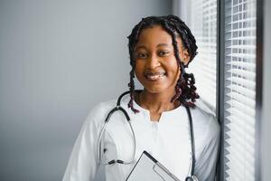 retrato confiante africano fêmea médico médico profissional escrevendo paciente notas isolado em hospital clínica corredor janelas fundo. positivo face expressão. foto