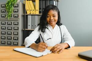 retrato confiante africano fêmea médico médico profissional escrevendo paciente notas isolado em hospital clínica corredor janelas fundo. positivo face expressão. foto