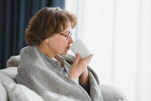 conforto relaxado Senior velho idosos mulher avó bebendo quente bebida chá café às casa olhando às Câmera dentro a vivo sala. pensão conceito. foto